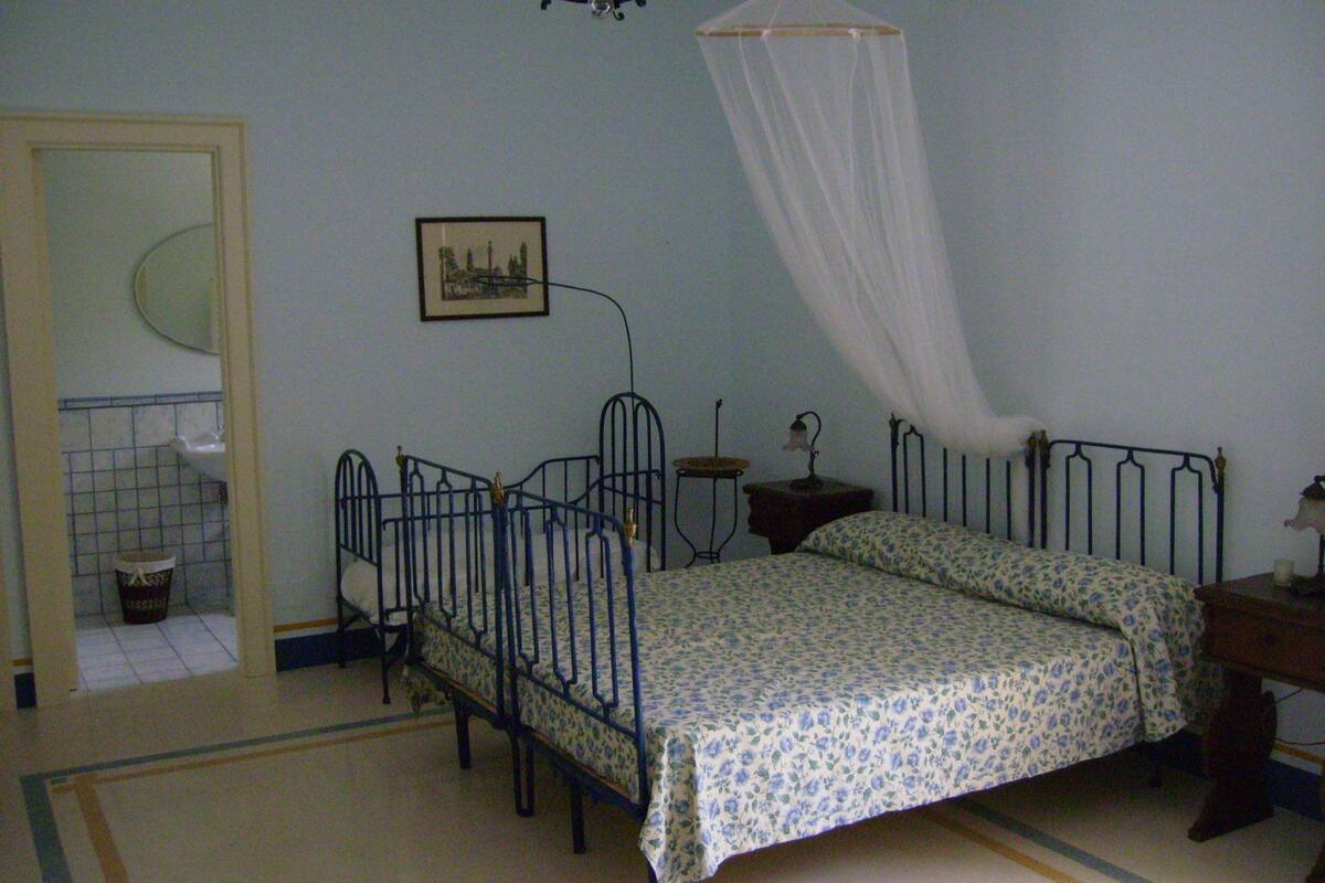 Une chambre avec un lit simple, une table de chevet, une commode, une lampe, un tableau au mur et une fenêtre avec un rideau.