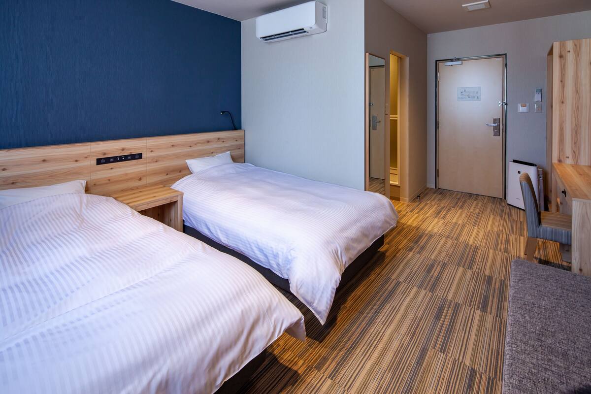 Habitación de hotel con dos camas, cabecera de madera, piso alfombrado, equipo de aire acondicionado y escritorio de madera.