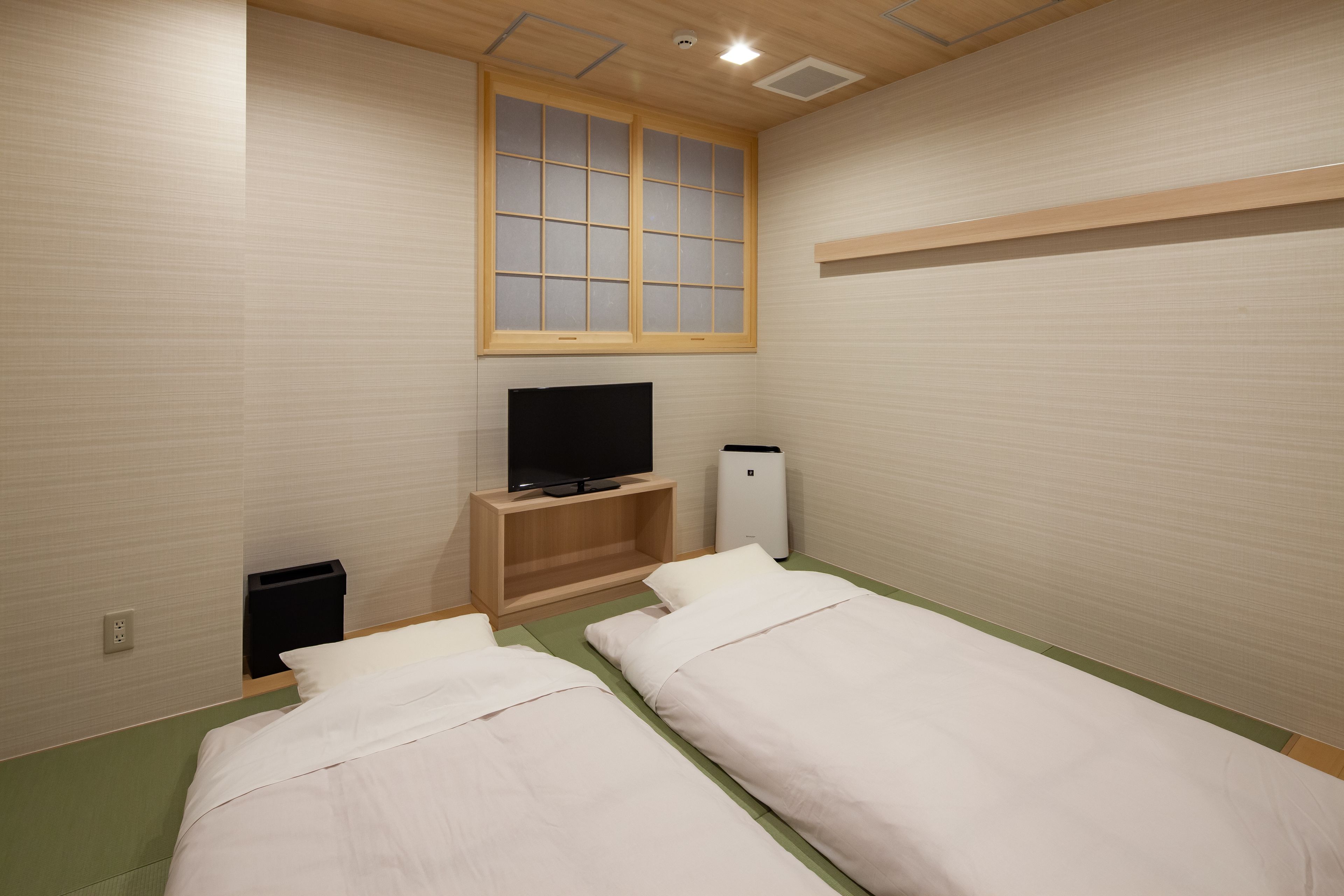 A room with two beds, a television on a wooden stand, a window with a grid pattern, and a white air conditioning unit.