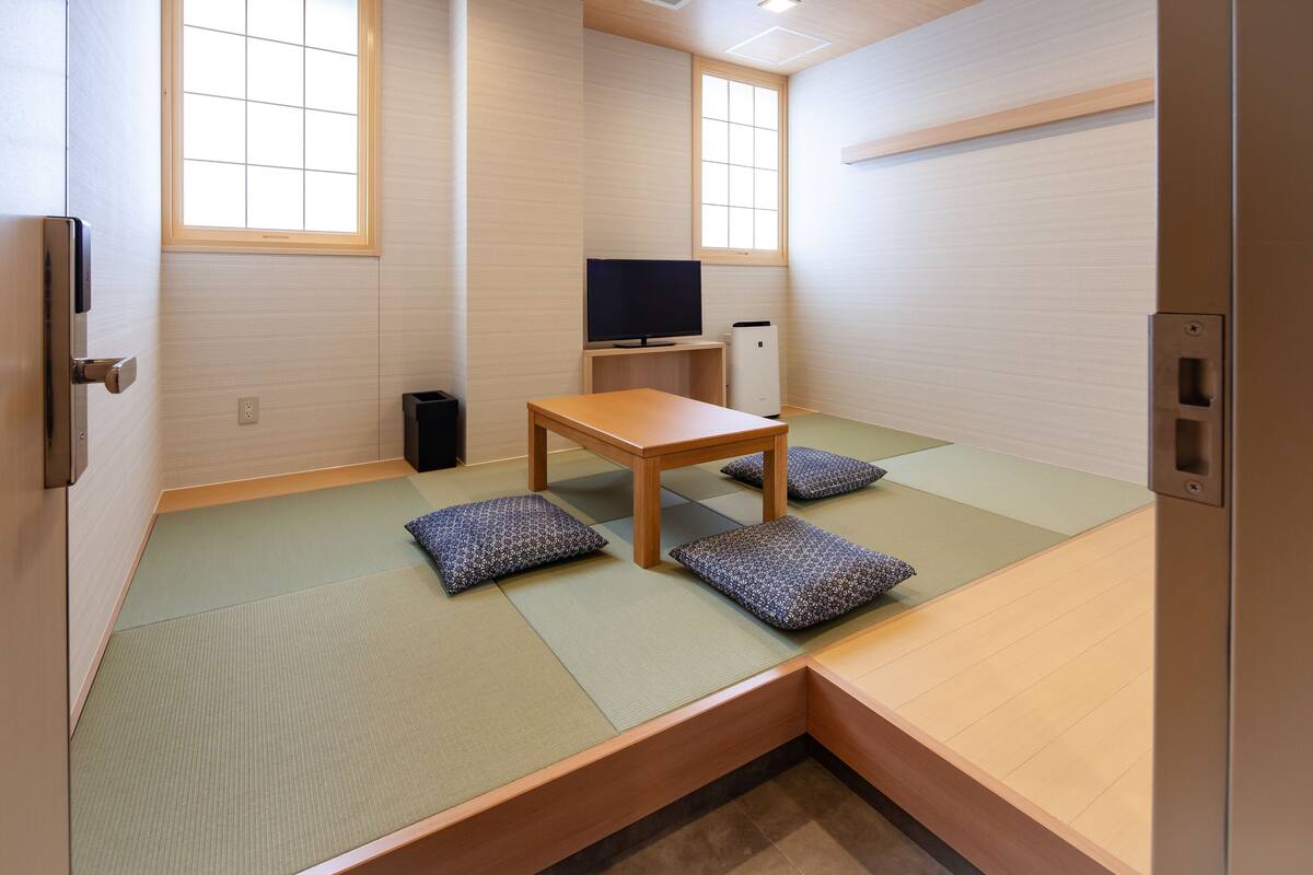 Una habitación con suelo de tatami, una mesa de madera baja, cojines, un televisor y un mueble pequeño.