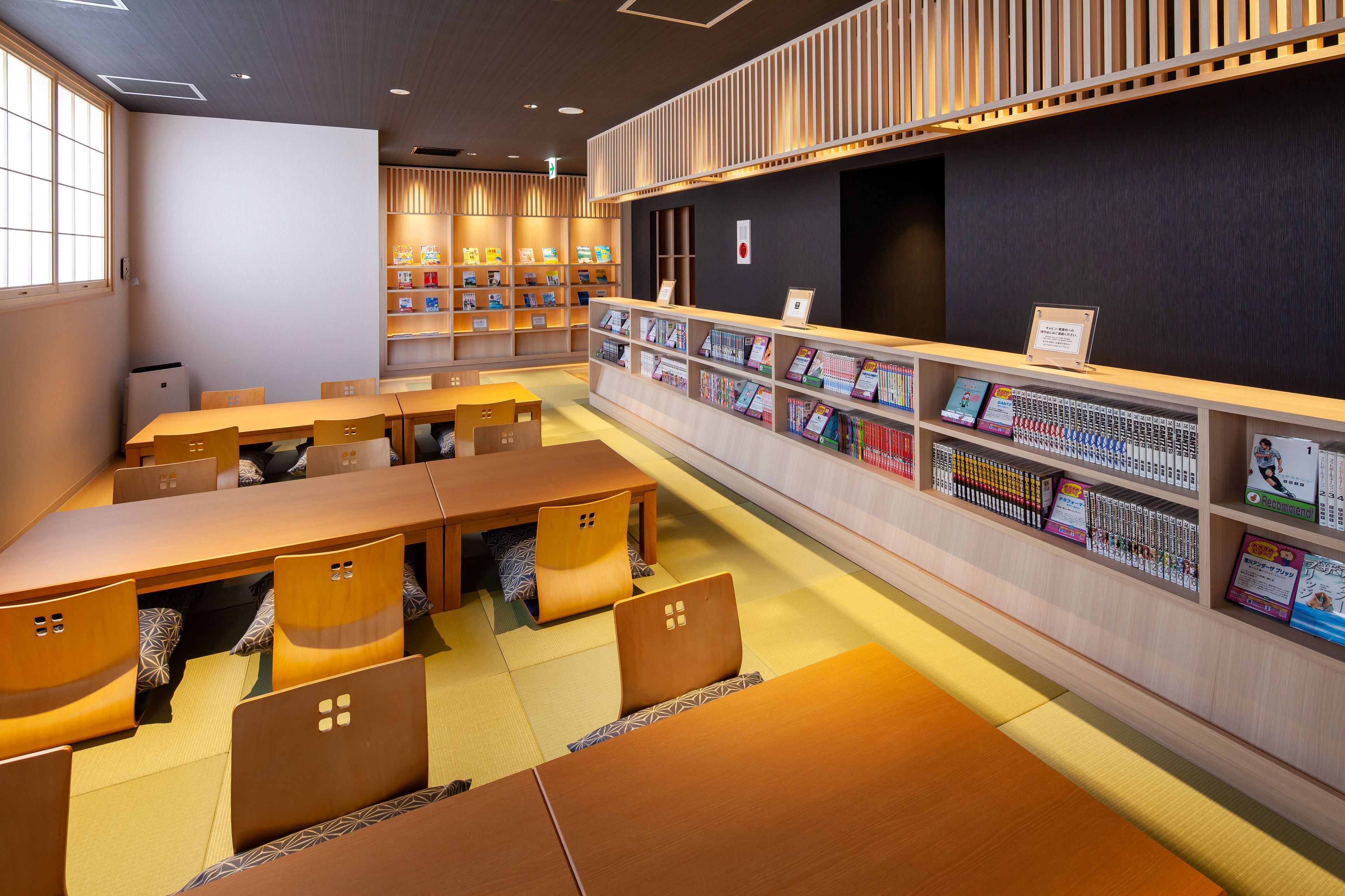 Una biblioteca con mesas y sillas de madera, estanterías y una pared oscura con un letrero rojo de salida de emergencia.
