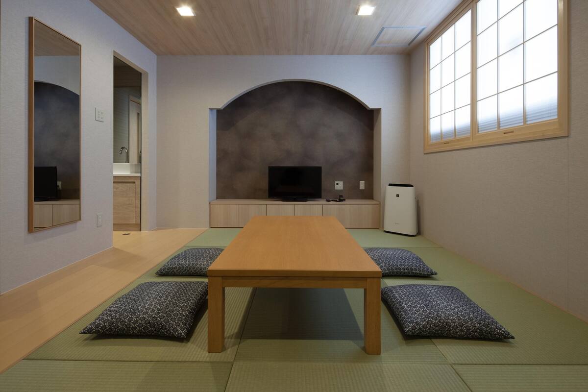 Una habitación con esteras de tatami, una mesa de madera, cojines, un televisor y un refrigerador pequeño.