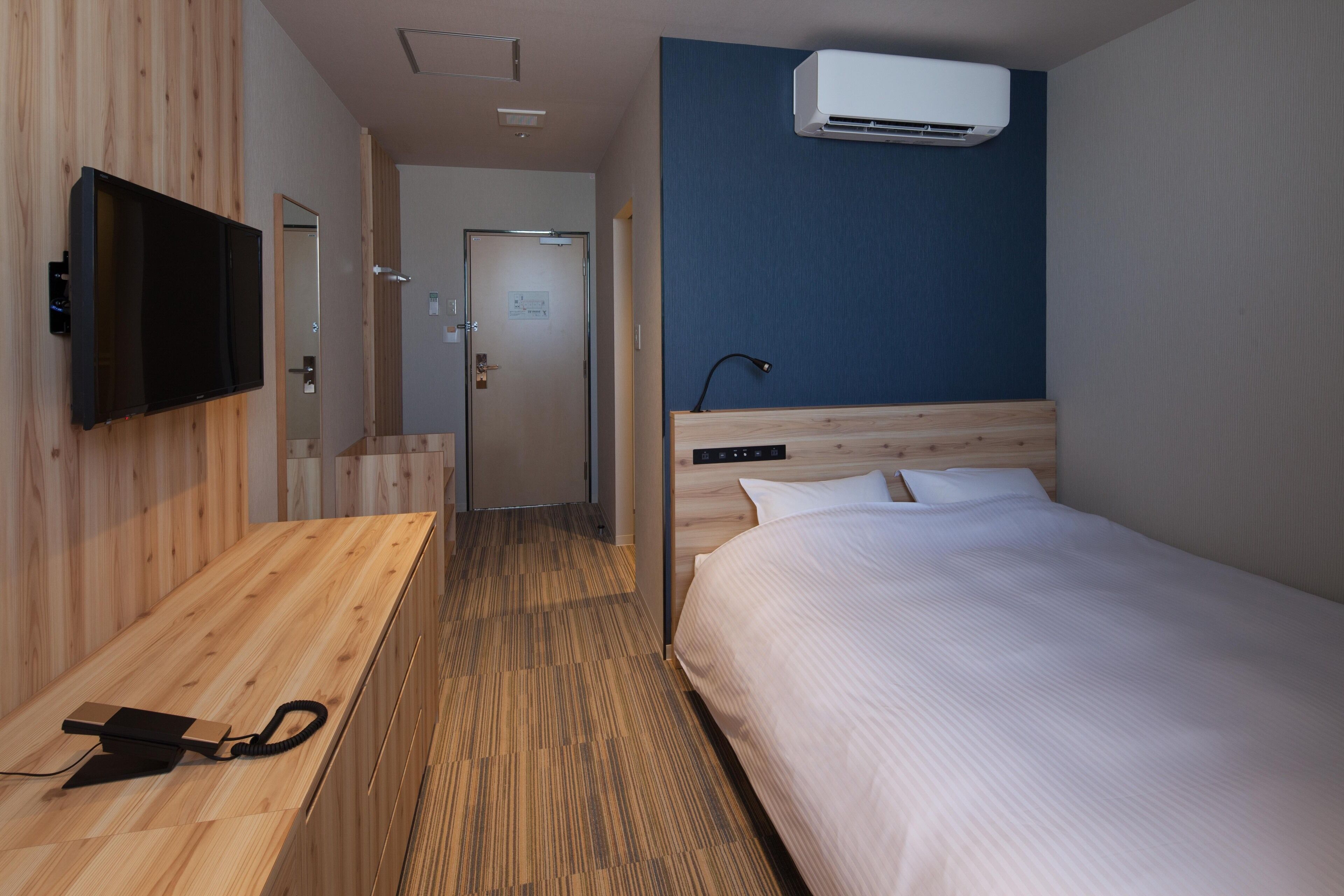 Hotellihuone, jossa on puinen työpöytä, sänky, seinälle asennettu televisio ja ilmastointilaite.