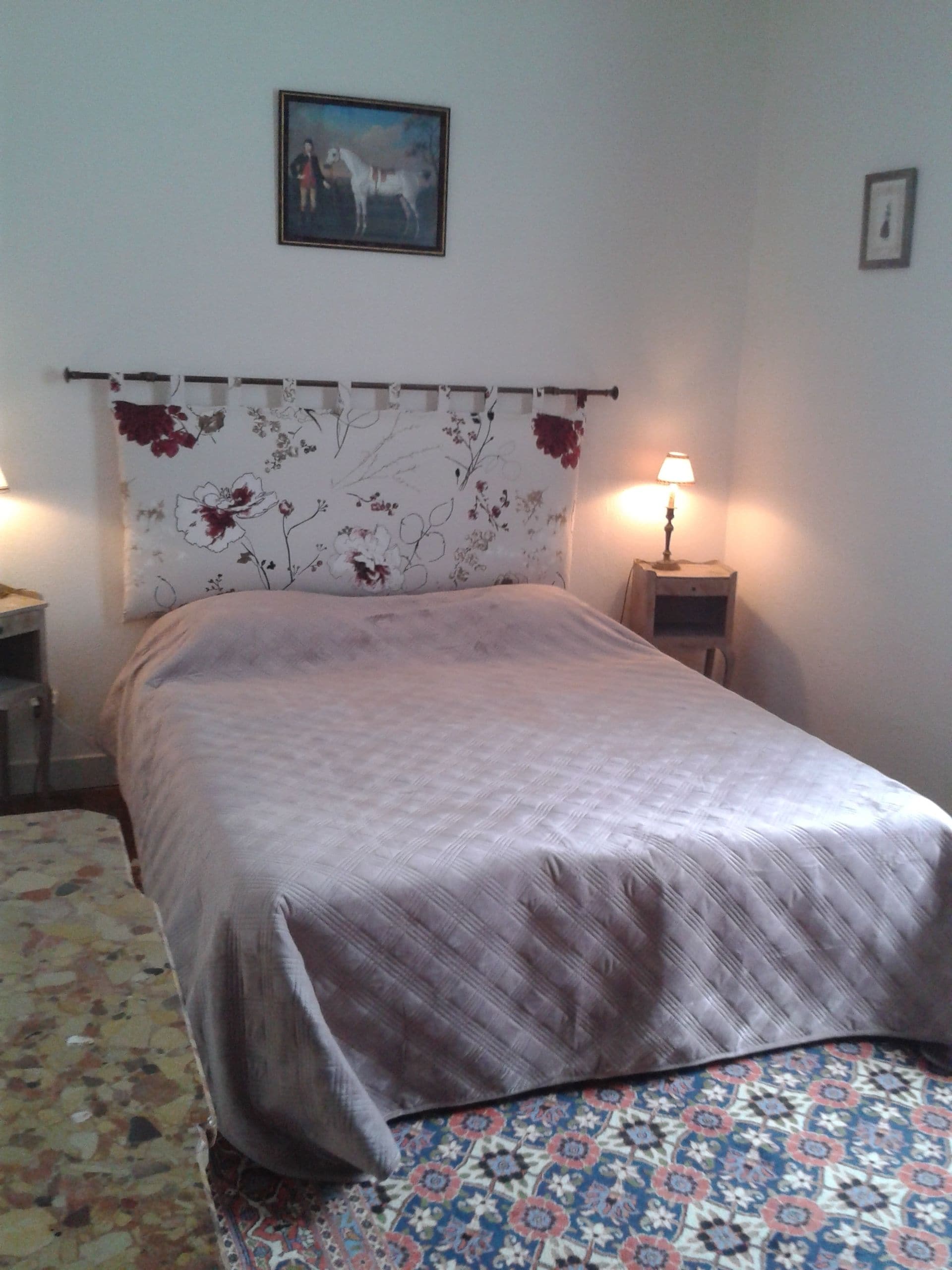 A bedroom with a bed, a nightstand, a lamp, a floral curtain, and a framed picture on the wall.