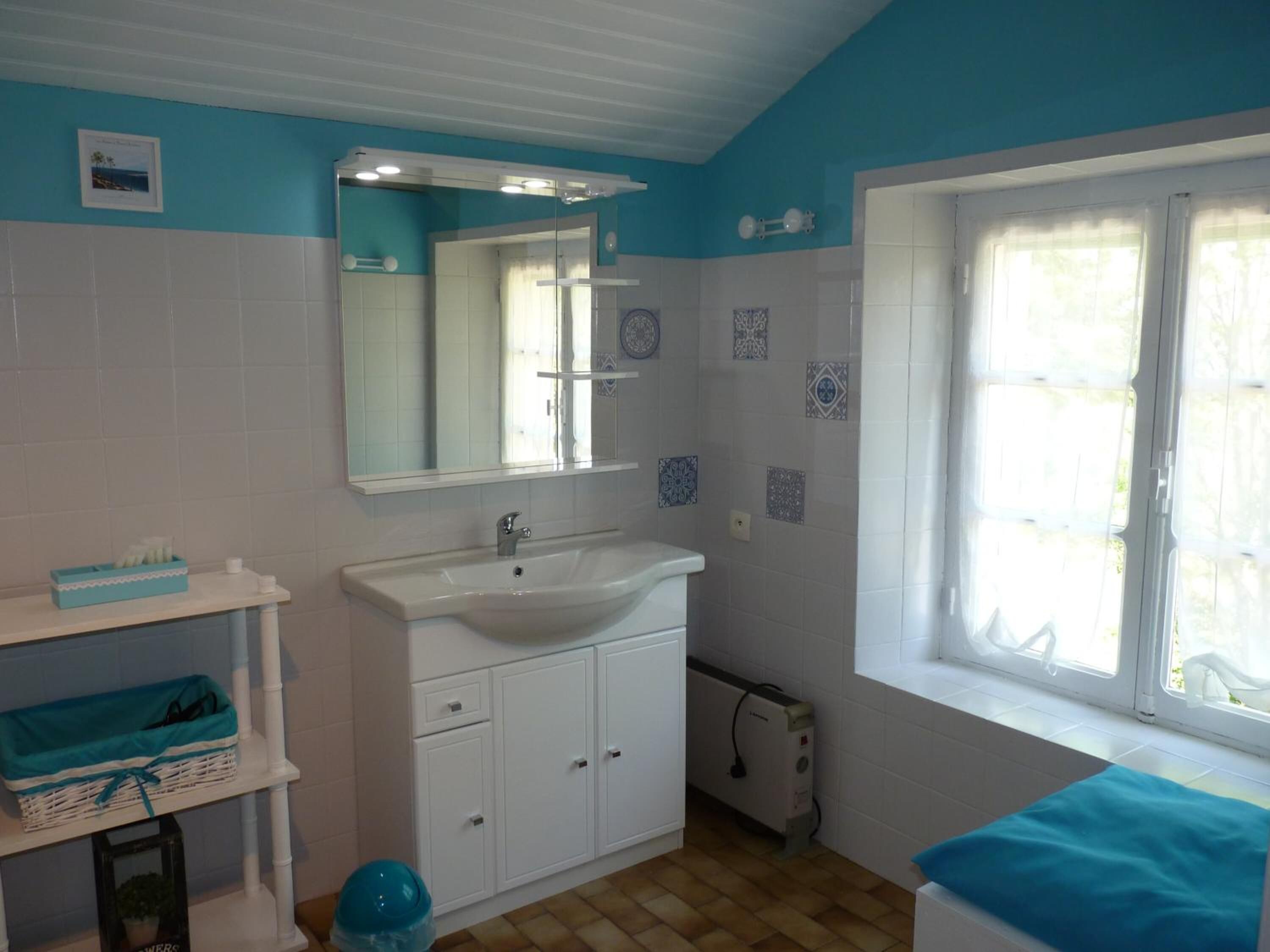 A bathroom with a white sink, a mirror, and a window with white curtains. There are decorative tiles on the wall and a small shelf with a blue basket.