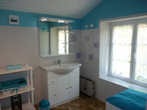 Un baño con un lavamanos blanco, un espejo y una ventana con cortinas blancas. Hay azulejos decorativos en la pared y un estante pequeño con una cesta azul.
