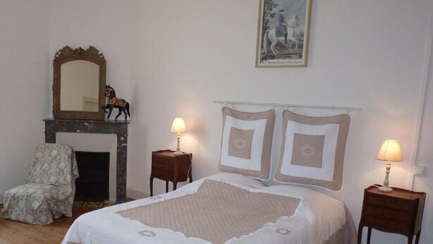 Een slaapkamer met een groot bed, een open haard, twee nachtkastjes met lampen, een stoel en een lijst aan de muur.