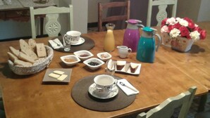 Een eettafel gedekt met brood, toevoegingen, een koffiepot, een karaf en een vaas bloemen.