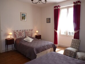 Un dormitorio con cama, dos mesitas de noche, una silla, un ventanal con cortinas y un candelabro.