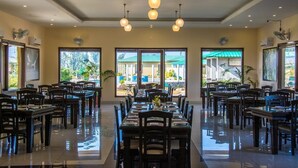 A spacious dining area with multiple tables and chairs, large windows offering views of the outdoors, and ceiling lights. - Corbett The Baagh Spa & Resort (Ramnagar)
