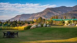 A golf course with a clubhouse, a dining area with chairs, and a view of mountains in the background. - Corbett The Baagh Spa & Resort (Ramnagar)