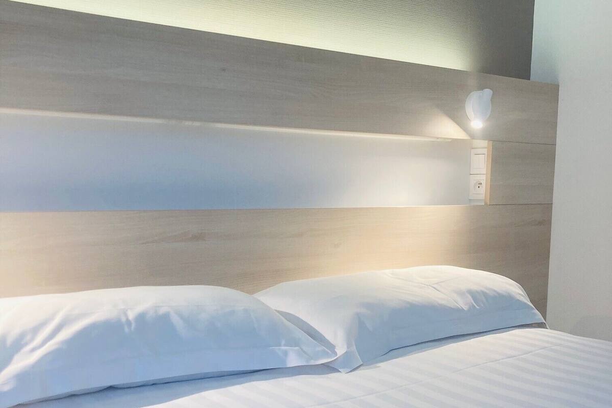 Una cama bien hecha con sábanas y almohadas blancas, cabecera de madera y una lámpara fijada a la pared.