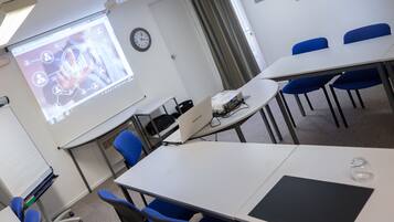 Un salón de conferencias con una pantalla de proyecciones, un reloj y sillas azules alrededor de una mesa blanca.