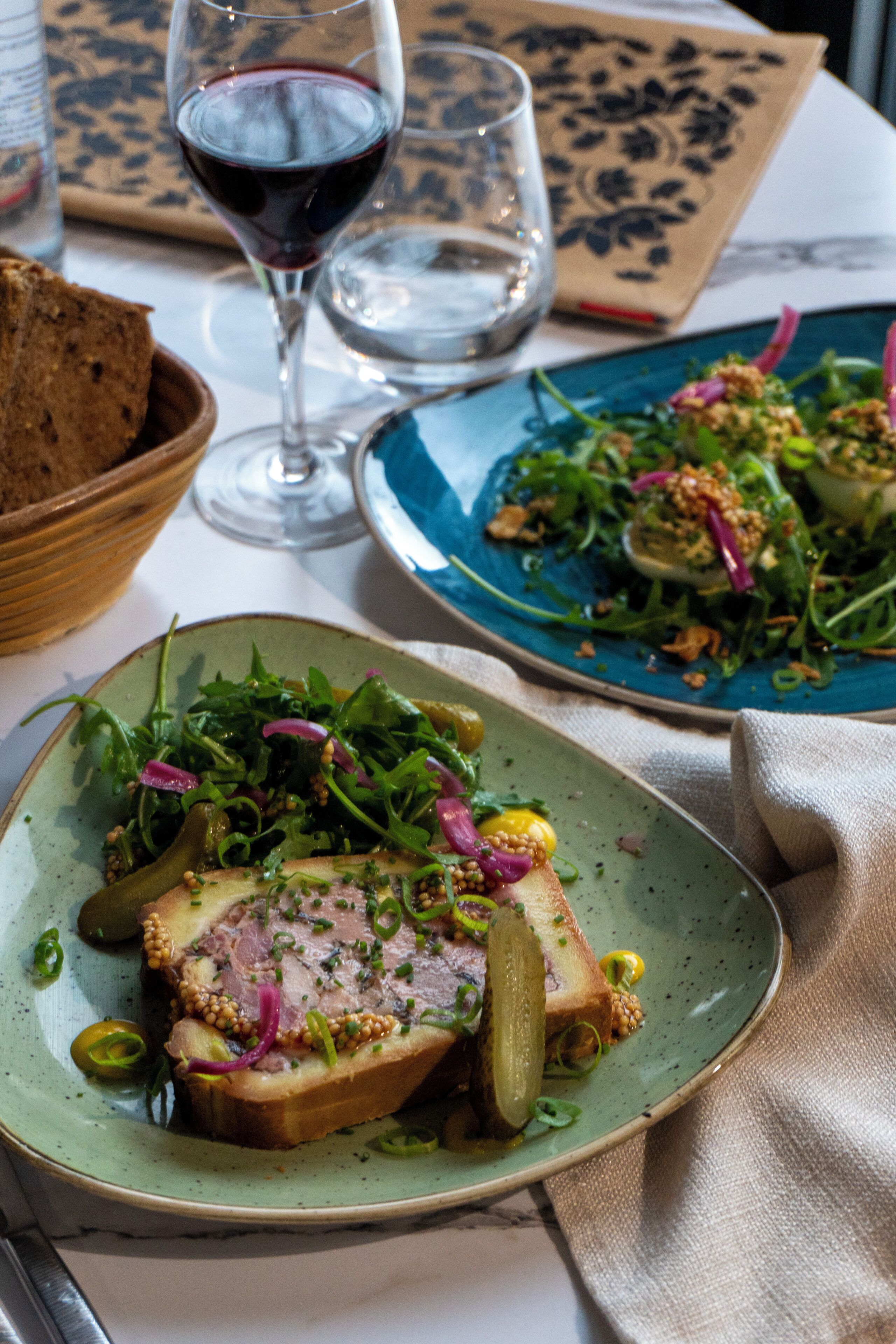 A dining table with plates of food, a glass of red wine, and a bowl of bread.