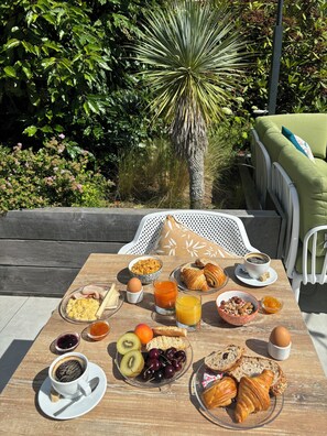 An outdoor breakfast table with a variety of foods and drinks, surrounded by lush greenery and a palm tree. - Hôtel Restaurant L’Eskemm *** St Brieuc-Trégueux (Tregueux)