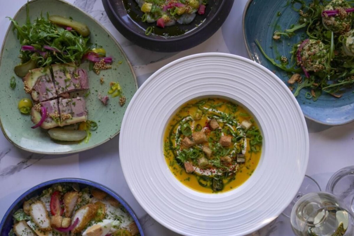 Eine Auswahl an Gerichten auf einem Tisch, darunter eine Suppe mit Croutons und Grünzeug, ein Teller mit geschnittenem Fleisch und Gemüse sowie eine Schüssel mit grüner Soße.