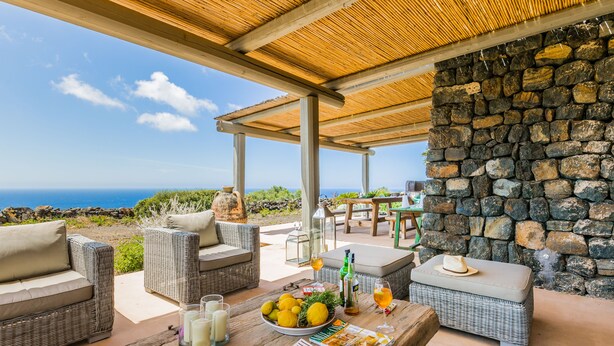 An outdoor seating area with wicker furniture, a stone wall, and a view of the ocean. - Wp Relais Dammusi Posidonia (pantelleria)