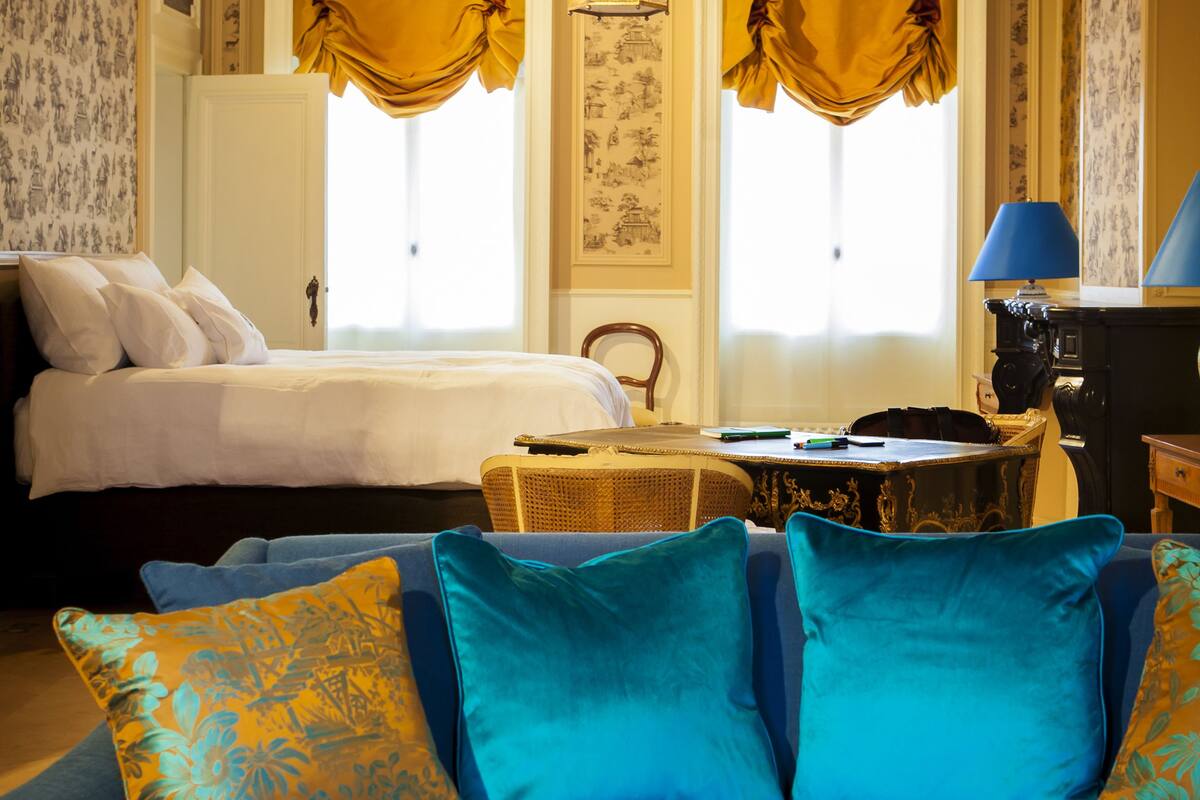 A hotel room with a blue sofa, two pillows, a bed, a table with two wine glasses and a vase with a rose, and two windows with yellow curtains.