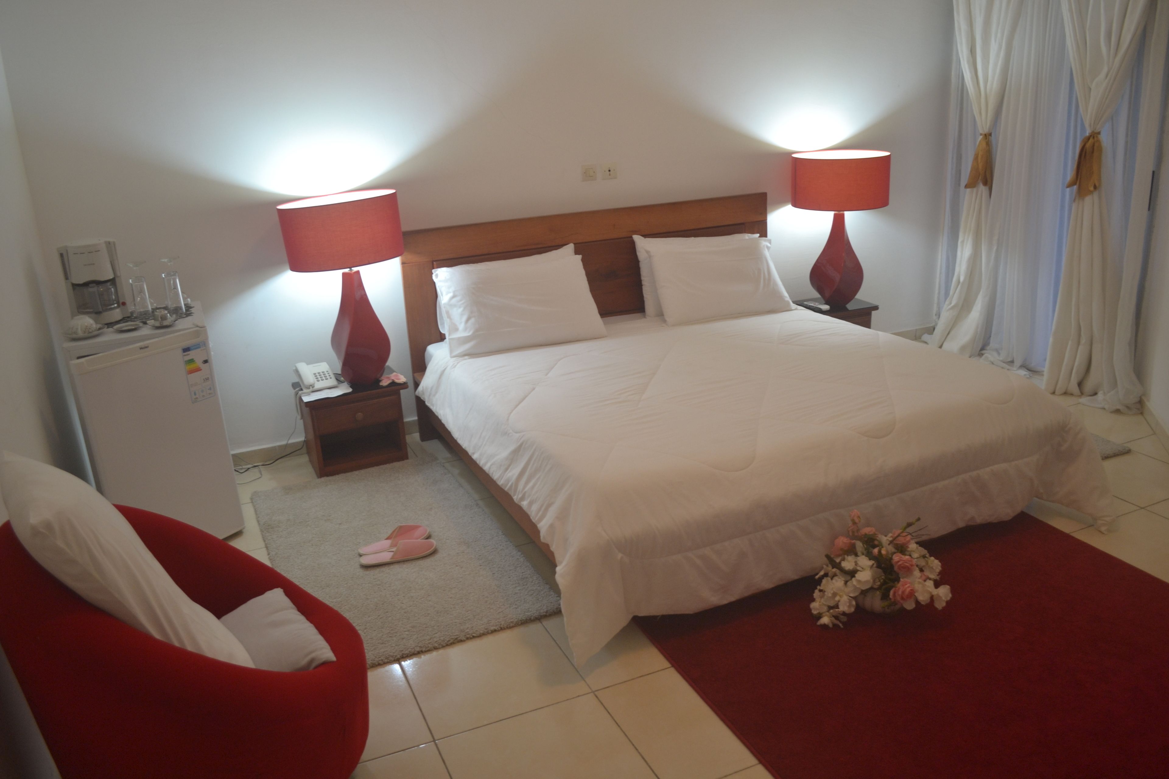 A hotel room with a large bed, two bedside lamps, a small desk with a phone, a red chair, and a red carpet.
