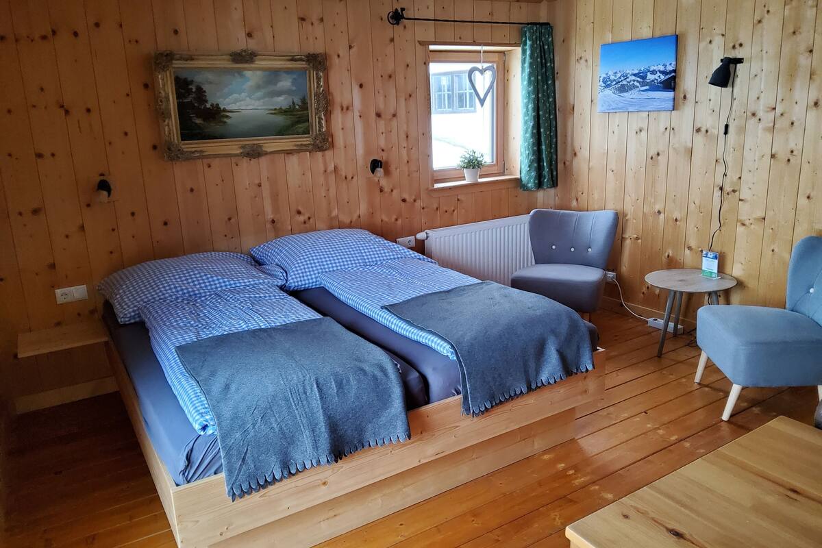 Puupaneloitu huone, jossa on sänky, tuoli, pieni pöytä, taulu ja ikkuna, jossa on verho.