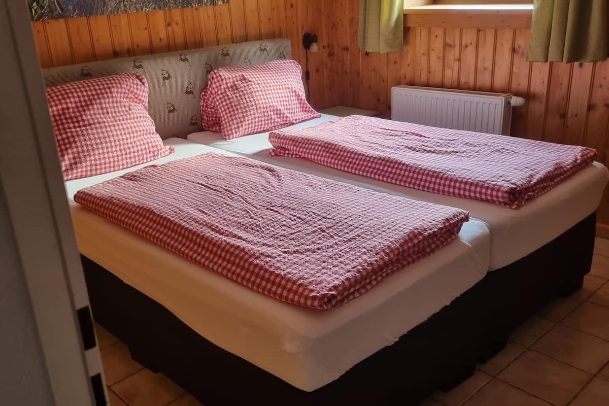 Kahden hengen huone, jossa on kaksi sänkyä. Jokaisessa sängyssä on ruutukuviollinen peitto ja tyynyjä. Huoneen seinät ovat puuta, ja siinä on ikkuna, jossa on verhot.