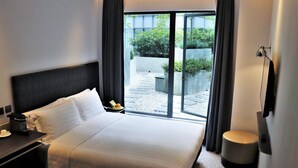 Habitación de hotel con una cama grande, una mesita de noche con lámpara, un taburete pequeño y vistas a un patio con plantas y un sendero.