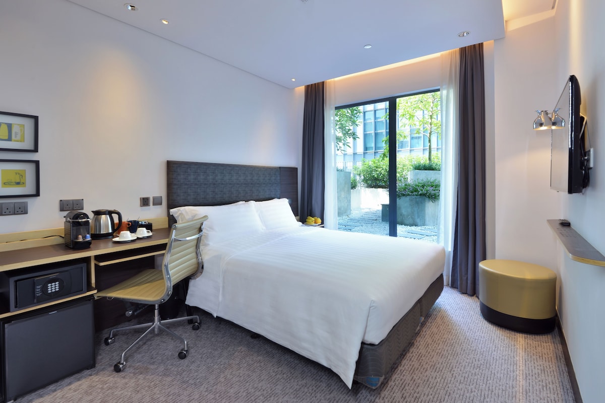 Une chambre d’hôtel moderne dotée d’un grand lit, d’un bureau avec une chaise, d’une petite kitchenette et offrant une vue sur la verdure environnante.
