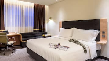 Hotellihuone, jossa on suuri sänky, työpöytä tuolilla, ikkuna verhoilla ja seinään kiinnitetty valaisin.