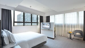 Habitación de hotel con una cama grande, un televisor de pantalla plana montado en la pared, una mesita redonda con frutero y un sillón gris.
