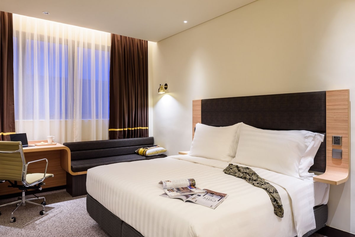 Une chambre d’hôtel avec un grand lit, un bureau avec une chaise, une fenêtre avec des rideaux et un luminaire fixé au mur.
