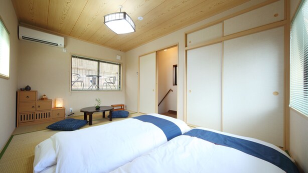 Een traditionele Japanse kamer met tatami vloeren, een lage tafel met kussens, een bed met wit en blauw beddengoed, en schuifdeuren.