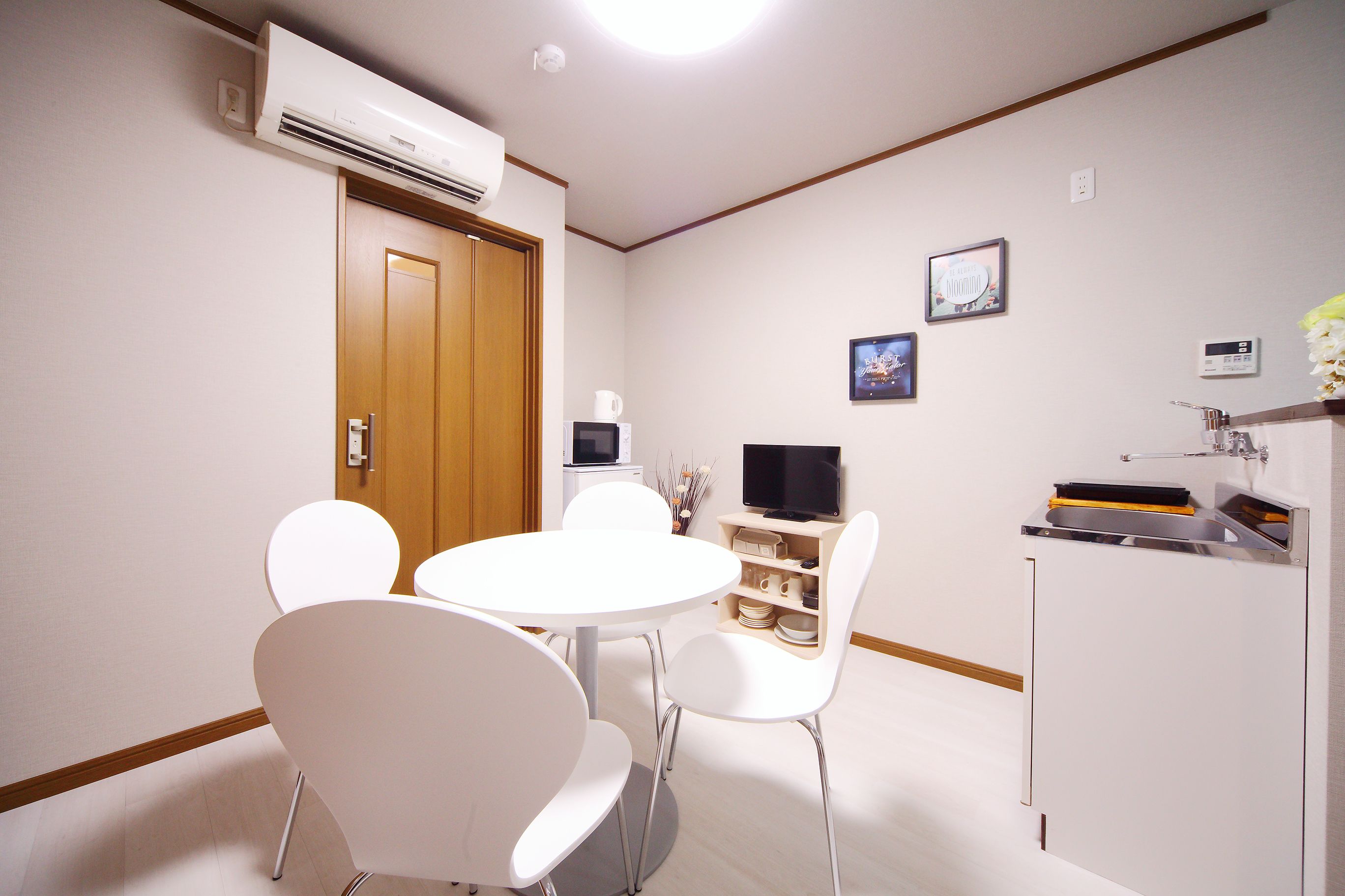 Ein modernes Zimmer mit rundem Tisch und weißen Stühlen, einer Holztür, einem an der Wand montierten Fernseher und einem kleinen Regal mit Dekoration.
