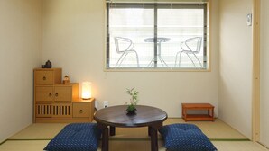 Een traditionele Japanse kamer met tatami vloeren, een lage houten tafel met een potplant en een raam met uitzicht op twee stoelen buiten.