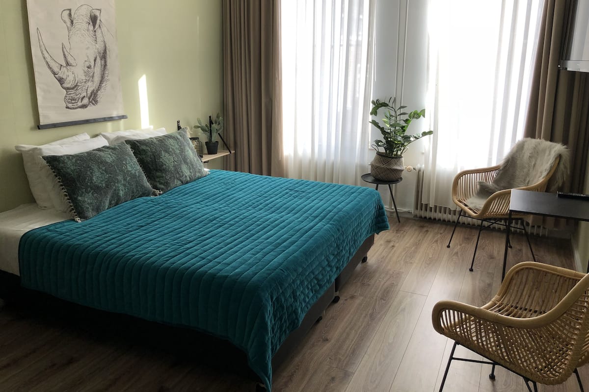 Une chambre à coucher comprenant un lit, deux oreillers, une chaise, une petite table, une plante en pot et une œuvre d’art accrochée au mur.