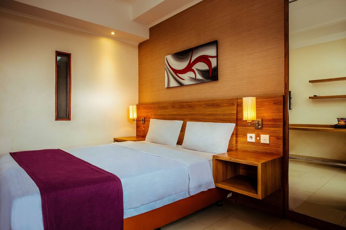 A hotel room with a large bed, wooden headboard, two bedside lamps, a wall-mounted artwork, and a small wooden shelf.