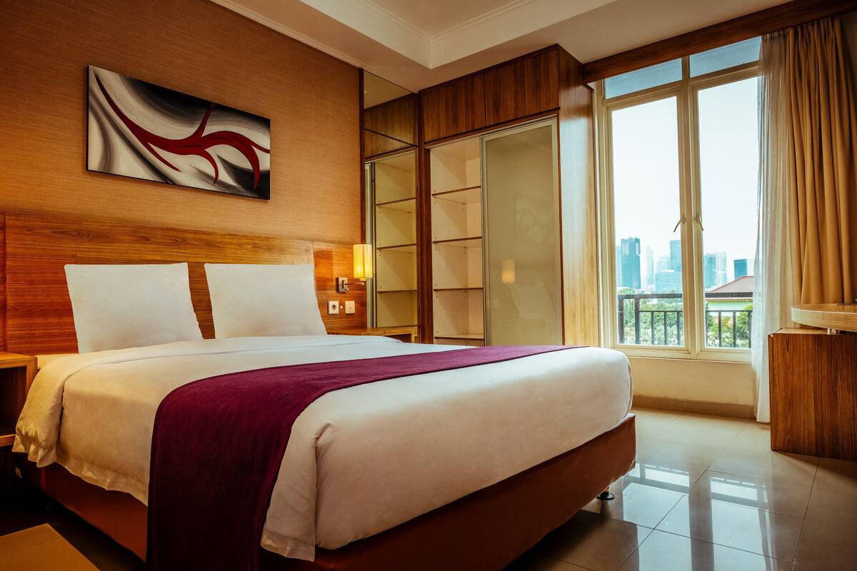 A hotel room with a large bed, a city view, a sliding door closet, and a modern abstract painting on the wall.