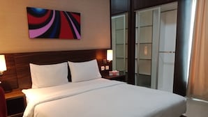 A hotel room with a large bed, two bedside lamps, a nightstand, a red chair, a wall-mounted abstract painting, and a sliding glass door leading to a balcony. - Marrakesh Inn (Jakarta)
