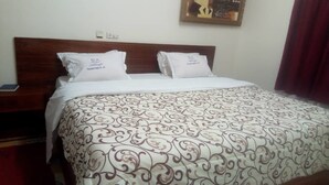 A neatly made bed with a patterned bedspread, two pillows with text, a nightstand with a phone, and a painting on the wall. - Hotel Aladje (Abidjan)