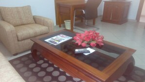 A living room with a sofa, a glass-top coffee table with a remote control and a magazine, a chair, a wooden cabinet, and a TV on top. - Hotel Aladje (Abidjan)