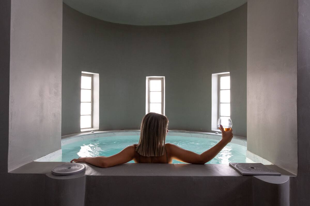 A person relaxing in a small, modern indoor pool with three windows, a glass of drink, and a scale on the edge.