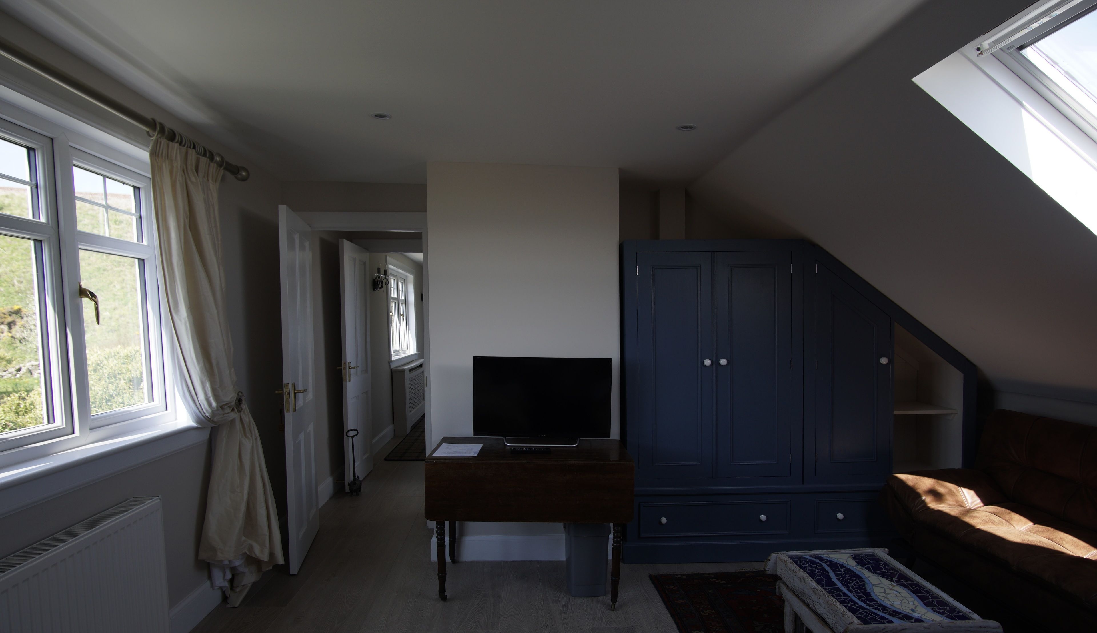 Una habitación con televisor sobre una mesa de madera, un armario azul oscuro, una ventana con cortinas y vista a zonas verdes.