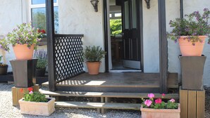 Un porche de madera con escalones que conducen a una puerta, flanqueado por plantas en macetas y una valla celosíaca.