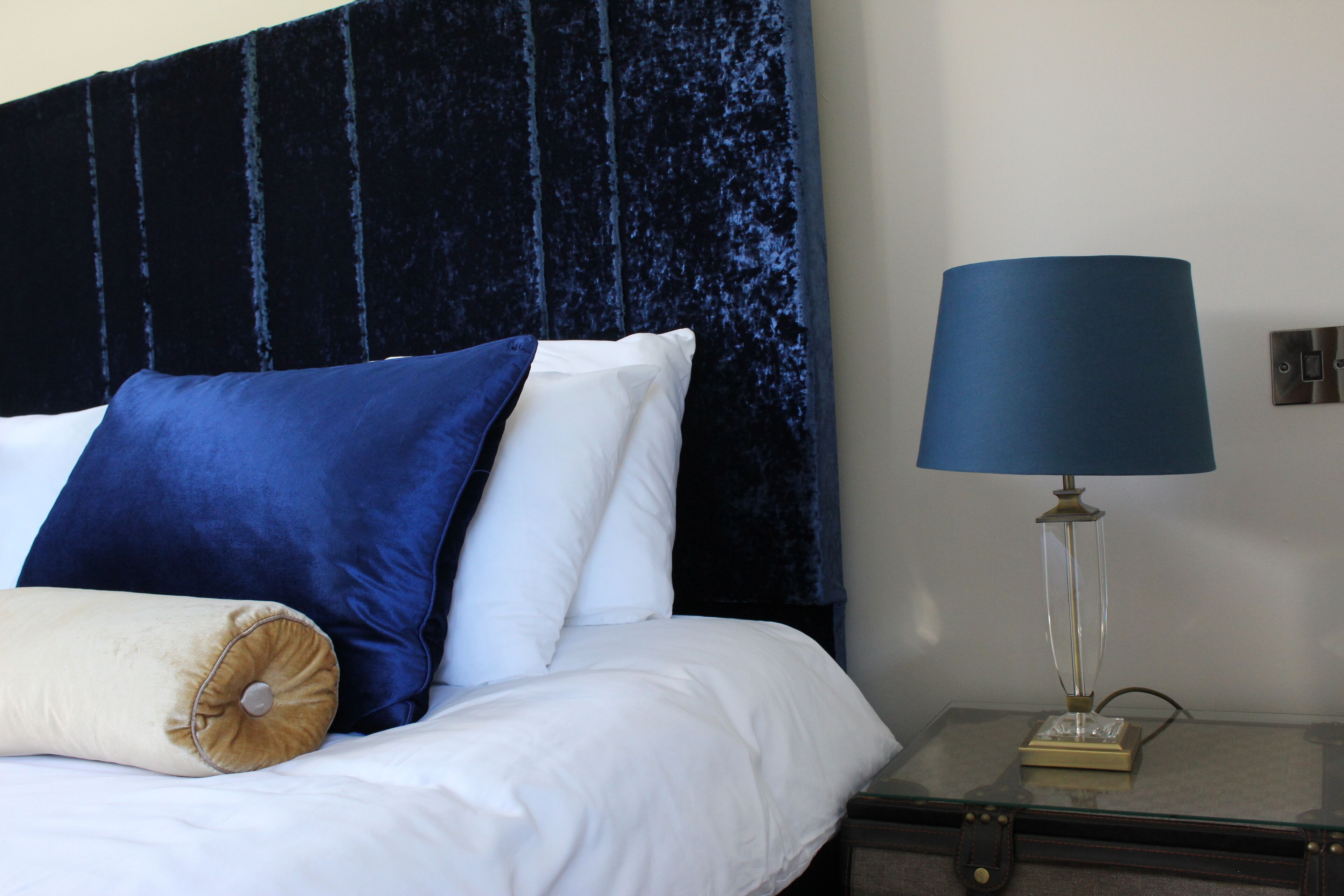 Una cama con ropa de cama blanca y azul, una lámpara azul sobre una mesita de noche y un cabecero azul oscuro.