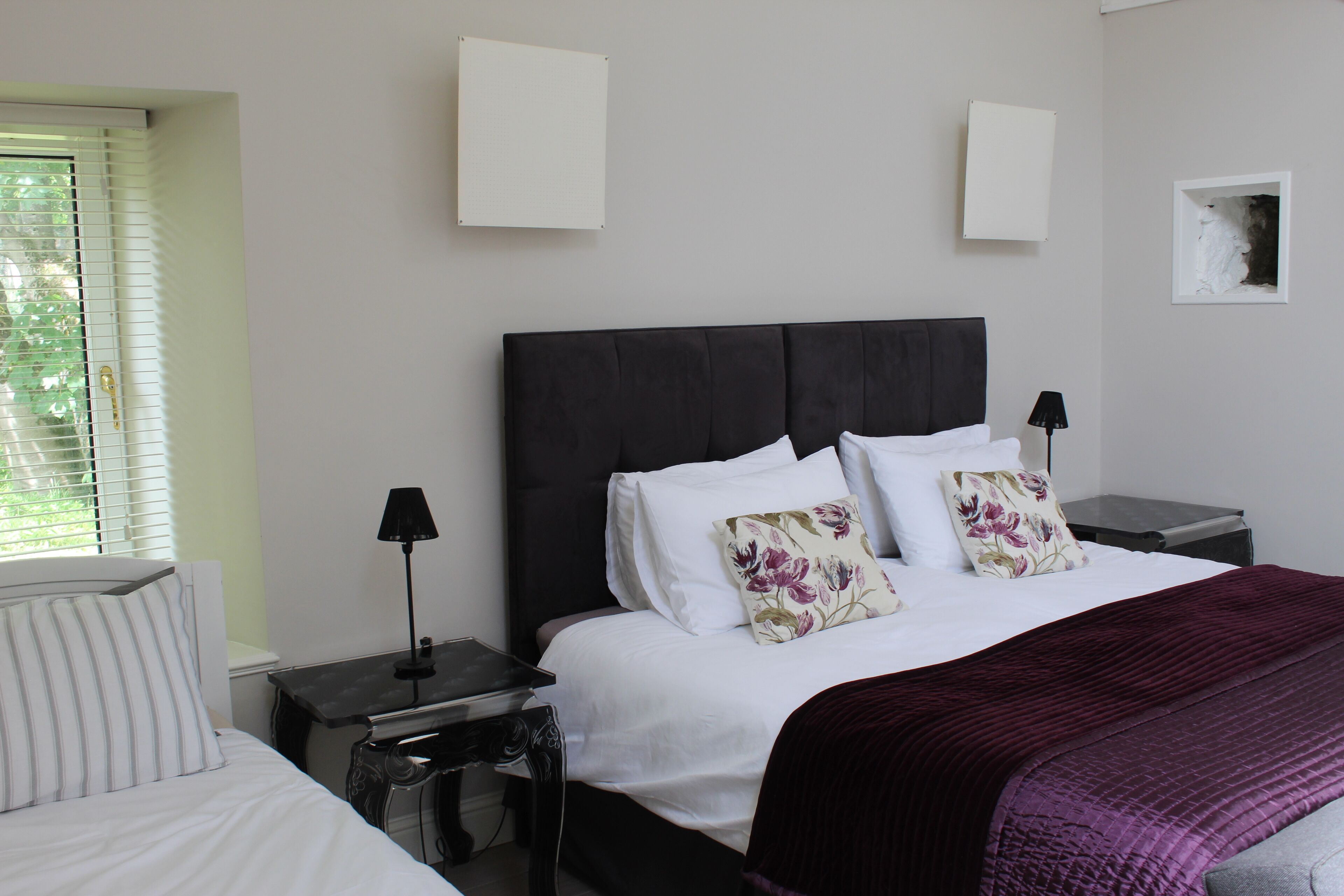 A bedroom with a large bed, two bedside tables, a window with blinds, and a wall-mounted artwork.