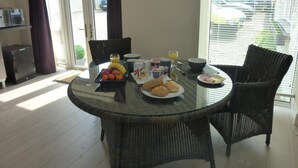 Un comedor con una mesa de vidrio rodeada de sillas de mimbre, una bandeja de desayuno con frutas, pan y bebidas, y vistas a través de un ventanal.