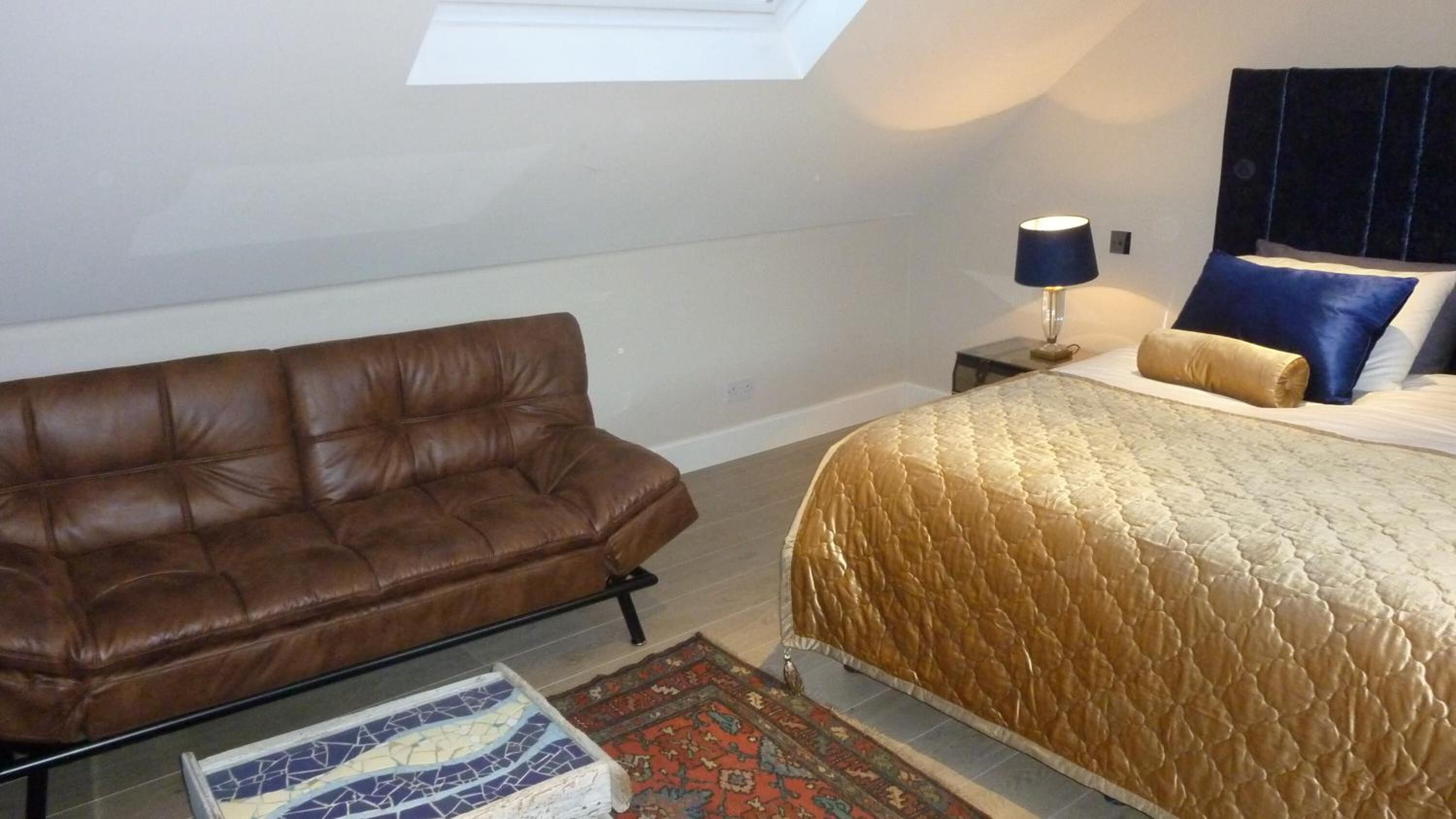 A bedroom with a brown leather sofa, a bed with a gold quilt, a nightstand with a lamp, and a patterned rug.