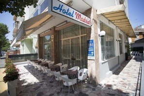 A hotel exterior with a sign reading "Hotel Madrid," outdoor seating with white chairs and patterned cushions, and a stone-paved entrance. - Hotel Madrid (Cattolica)