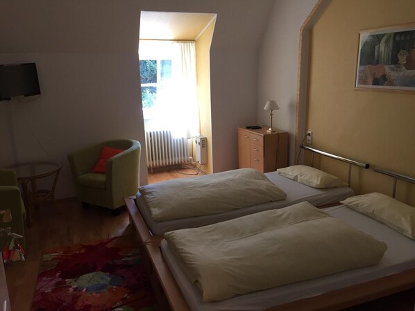 Habitación de hotel con dos camas, un sillón verde, una mesita y un radiador, además de un cuadro en la pared.