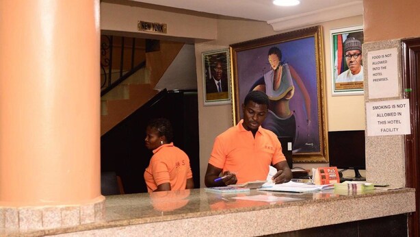 A hotel reception desk with staff members. There are framed portraits on the wall, a sign prohibiting food and smoking, and a sign indicating the location as New York. - B & S Hotel (Lagos)