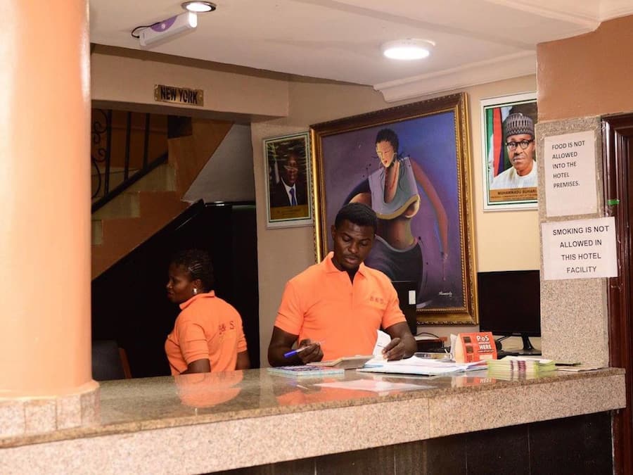 Un mostrador de recepción de hotel con personal. Hay retratos enmarcados en la pared, un cartel que prohíbe comer y fumar, y un letrero que indica la ubicación como Nueva York.